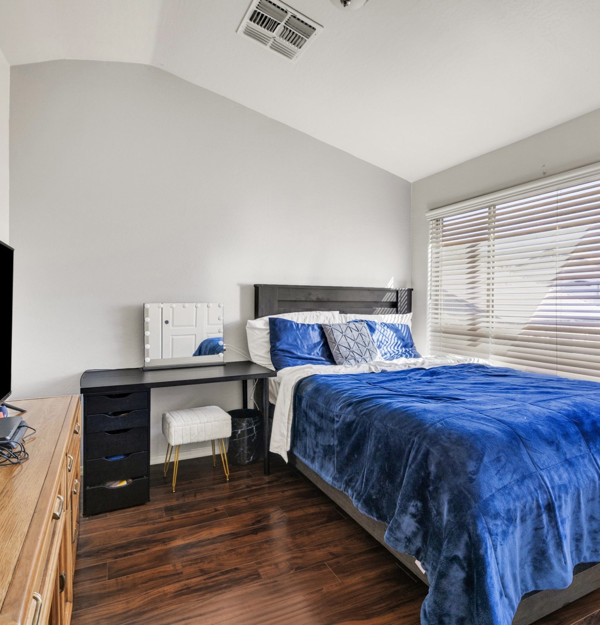 Interior of a spacious upstairs bedroom at 845 S Cerise in Mesa, AZ, featuring a high vaulted ceiling, dark wood flooring, a large window with blinds, and a black desk and bed frame with blue bedding in the gated La Fortina community.
