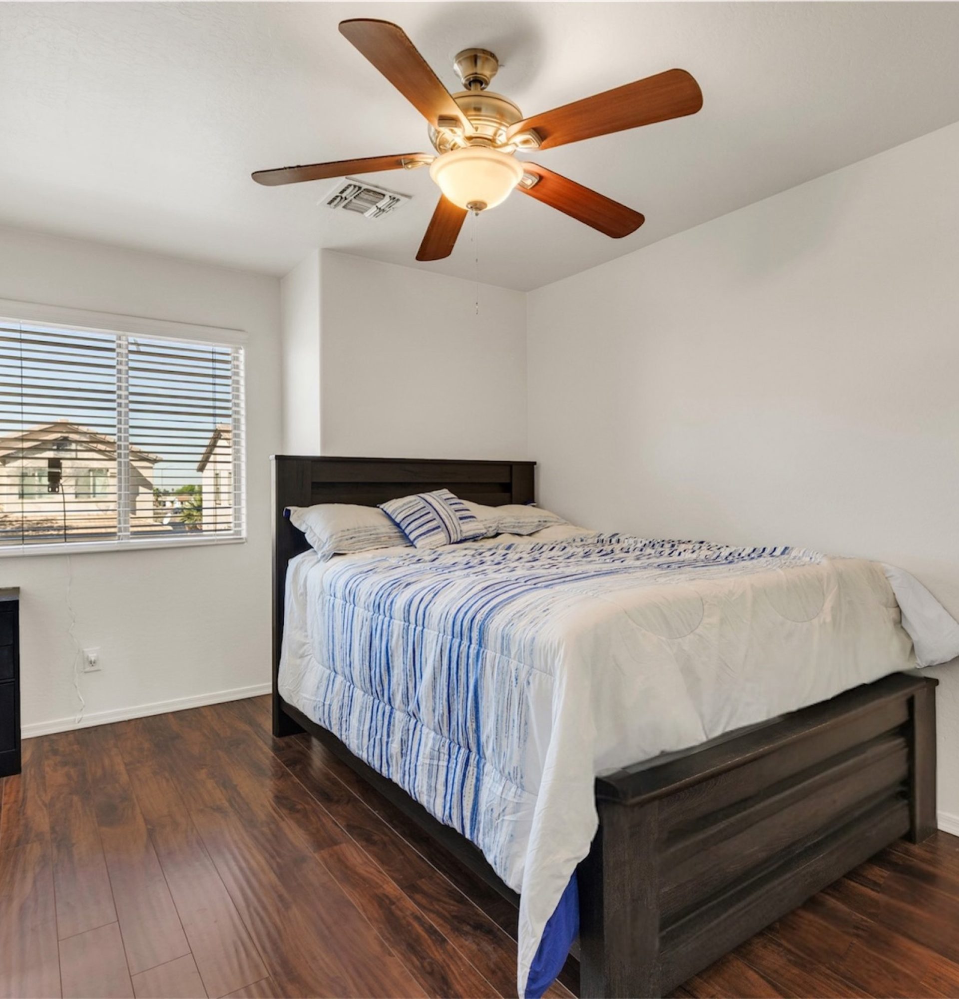 Interior of a secondary bedroom at 845 S Cerise in Mesa, AZ, featuring rich dark wood flooring, a queen-sized bed with blue and white bedding, a modern ceiling fan, and a large window with blinds in the gated La Fortina community.