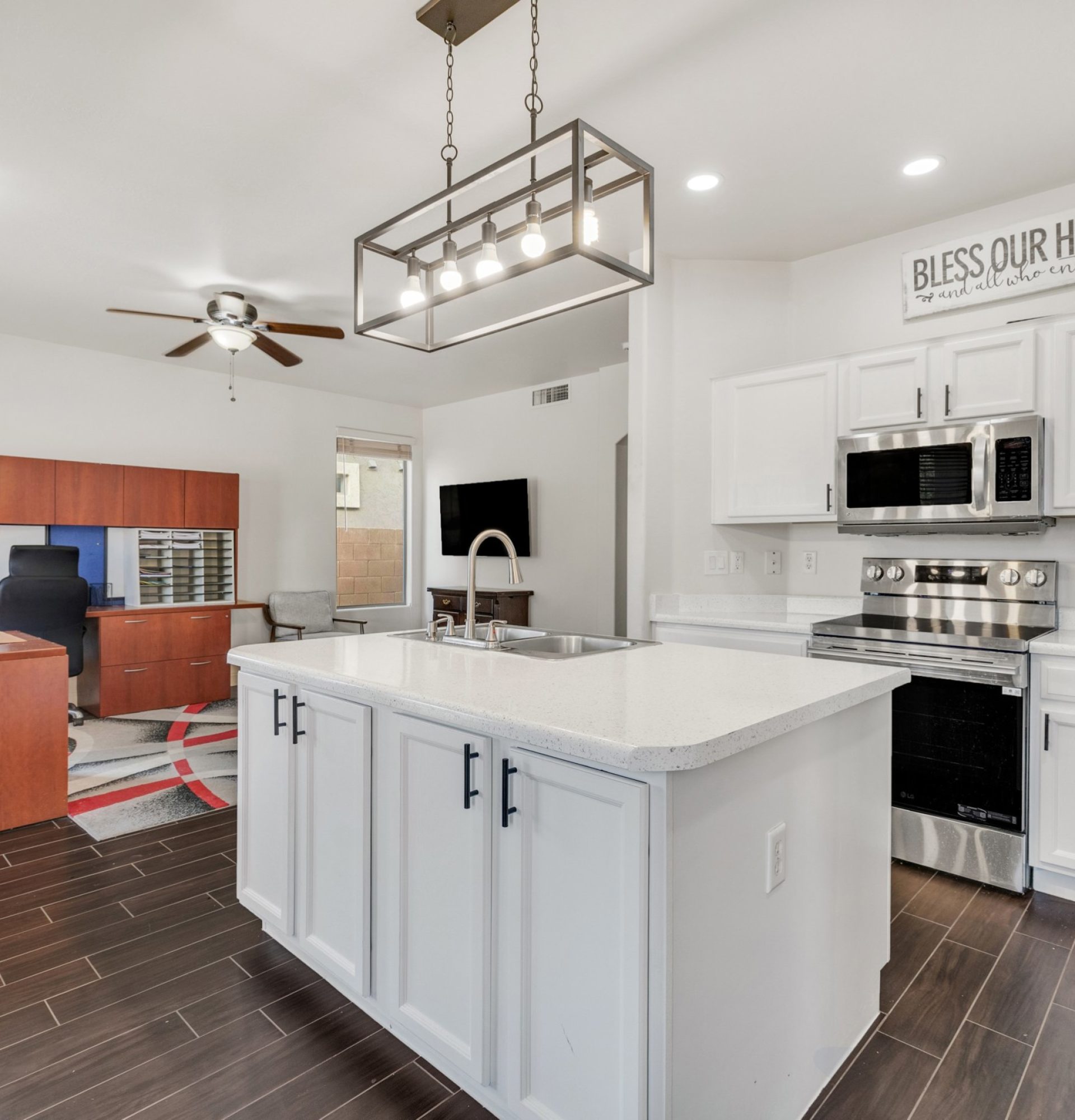 Wide-angle view of the open-concept kitchen and living area at 845 S Cerise in Mesa, AZ, featuring a white island, stainless steel appliances, dark wood-look tile, and a spacious area currently used as a home office setup in the gated La Fortina community.