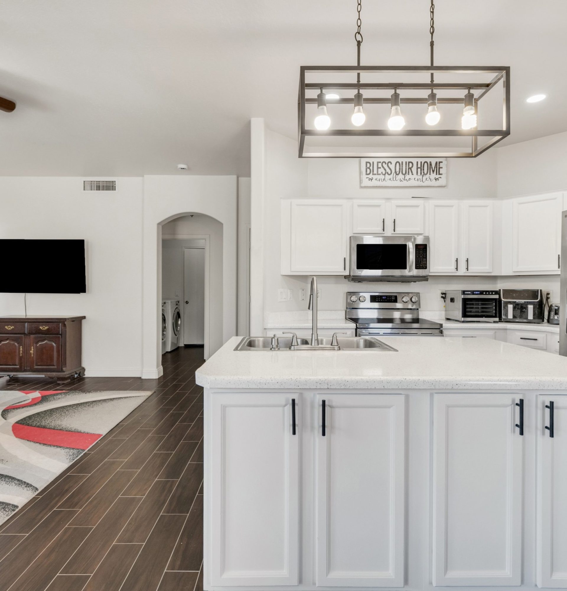 Modern kitchen interior at 845 S Cerise in Mesa, AZ, featuring a large white island with a sink, stainless steel appliances, white cabinetry with black hardware, a contemporary linear chandelier, and dark wood-look tile flooring.