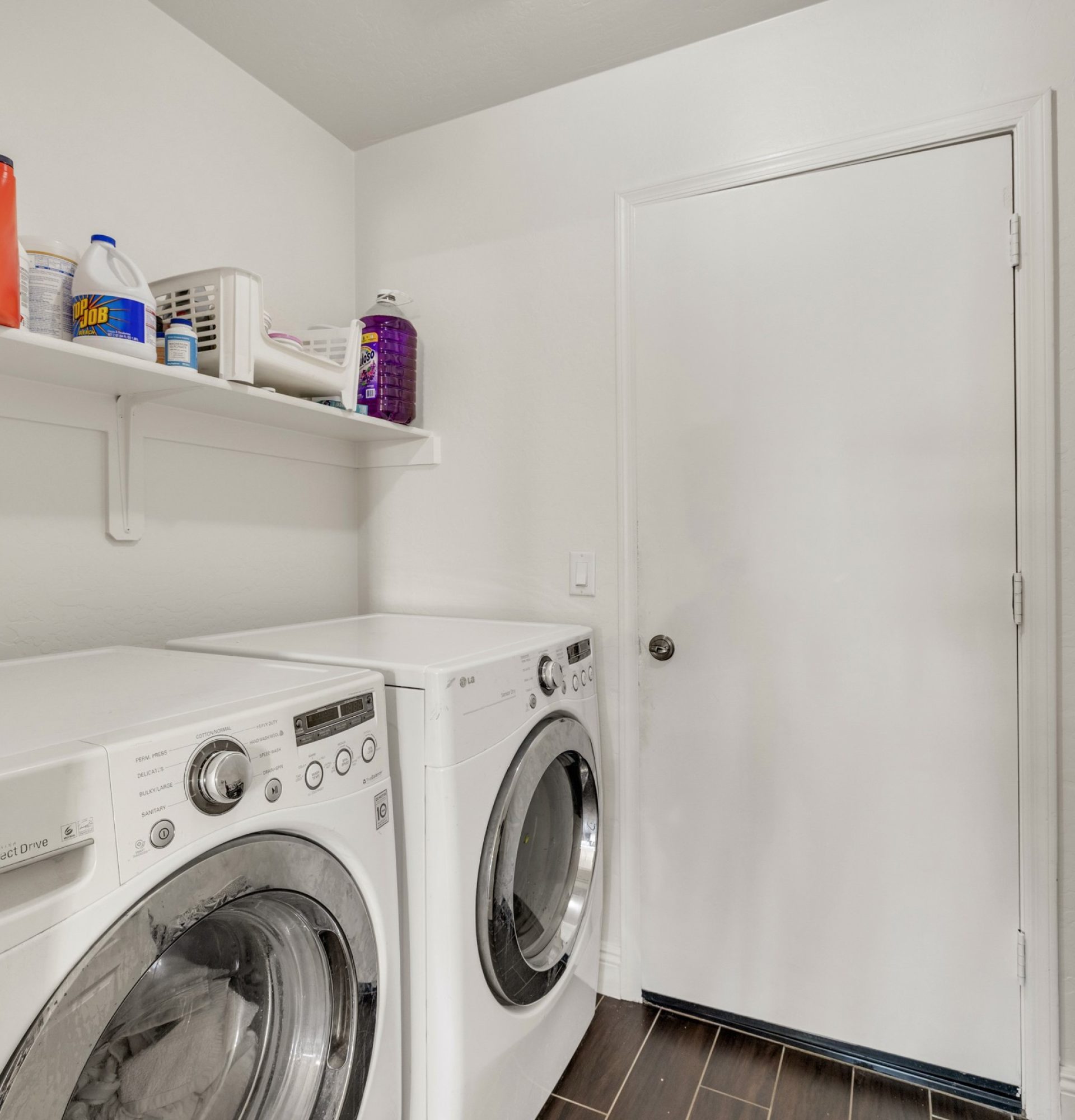 Interior of the laundry room at 845 S Cerise in Mesa, AZ, featuring white LG front-load washer and dryer units, a built-in storage shelf with supplies, neutral white walls, and dark wood-look tile flooring in the gated La Fortina community.