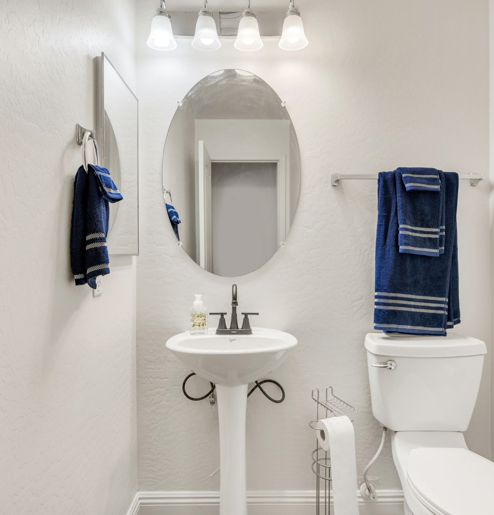 A bright, modern half-bathroom at 845 S Cerise, Mesa, AZ, featuring a white pedestal sink, oval mirror, four-light vanity fixture, and neutral white walls with blue towel accents in the La Fortina community.