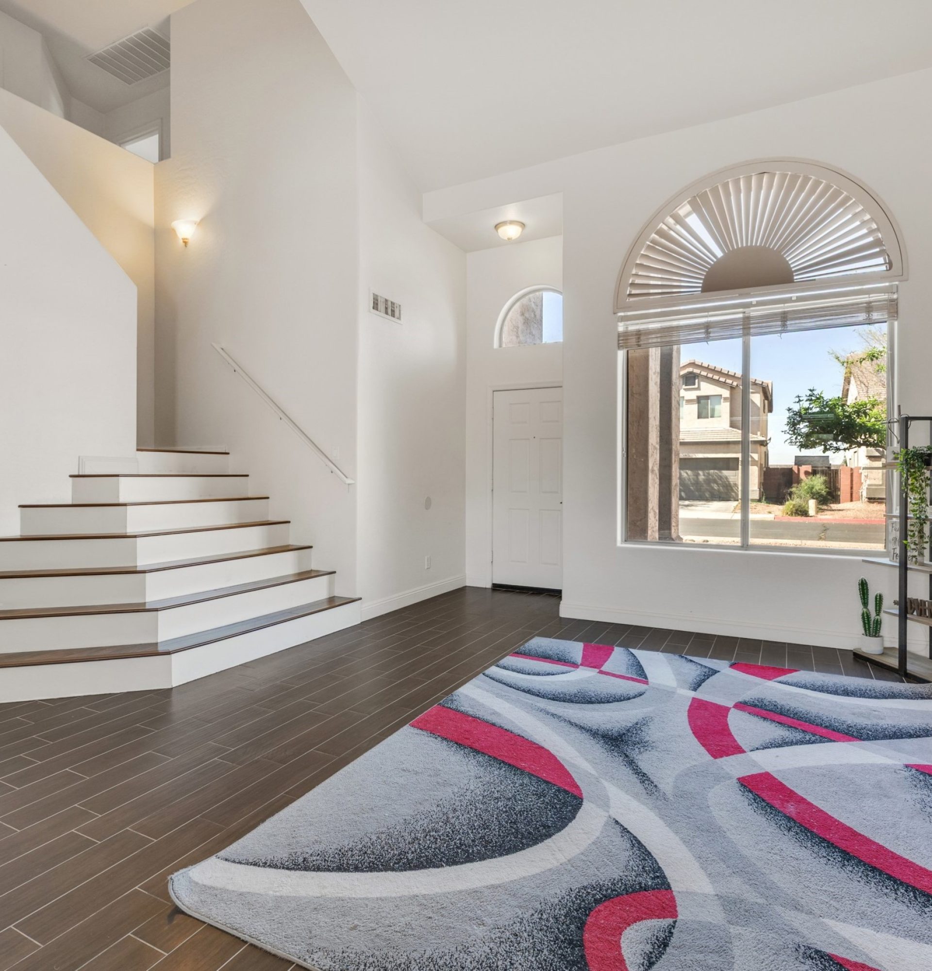 Interior view of the formal entryway and living area at 845 S Cerise in Mesa, AZ, featuring vaulted ceilings, wood-look tile flooring, a grand staircase with white risers, and a large arched window with a sunburst blind in the La Fortina community.
